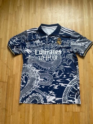 Real Madrid draktröja Adidas blå - Säljer en blå Real Madrid matchtröja från Adidas med ett fett drakmönster i vitt över hela tröjan. Tröjan har korta ärmar, krage och klubbmärke på bröstet. Materialet är troligtvis polyester, perfekt för fotboll eller träning.
