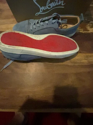 Blåa Louboutin sneakers med röd sula - Snygga blåa sneakers från Christian Louboutin i mocka med den ikoniska röda sulan. Skorna har lågt skaft, matchande blå skosnören och vita kanter runt sulan. Perfekta för dig som vill sticka ut med exklusiva detaljer och klassisk design.