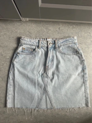 Ljusblå jeanskjol - Trendig ljusblå jeanskjol i kort modell med rå, fransig kant nedtill. Kjolen har klassiska fem fickor, bälteshällor och en snygg asymmetrisk söm framtill. Perfekt för en avslappnad och cool stil.