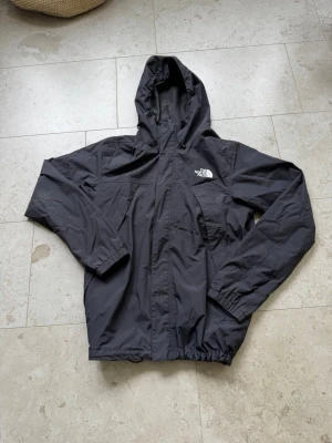 Svart vindjacka från The North Face - Snygg svart vindjacka från The North Face med huva och dragsko. Jackan har dragkedja framtill, elastiska muddar och logga på bröstet. Tillverkad i lätt och tåligt material som står emot vind. Perfekt för blåsiga dagar och outdoor-äventyr.