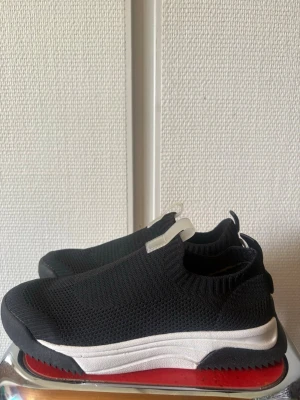 Svarta slip-on sneakers med röd sula - Snygga svarta slip-on sneakers med stickad ovandel och vit mellansula. Yttersulan har en cool röd detalj undertill och svart kant. Skorna har en sportig vibe och är superlätta att ta på tack vare elastisk resår och flärp bak. Perfekta för dig som gillar stilrena och bekväma sneakers.