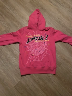 Spider hoodie / rosa  - Cool rosa hoodie med tryck av ett stort spindelnät och texten 'PINK!' framtill. Hoodien har huva, långa ärmar och ribbade muddar. På baksidan finns en gul stjärna och små färgglada detaljer. Perfekt för dig som gillar färg och statement-plagg.