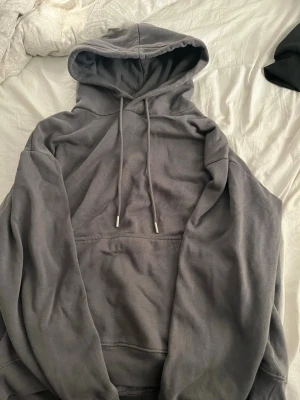 Mörkgrå hoodie med huva - En enkel och stilren mörkgrå hoodie från HM med stor huva och dragsnören. Hoodien har en klassisk känguruficka framtill och ribbade muddar vid ärmslut och nederkant. Perfekt för en avslappnad och bekväm look.