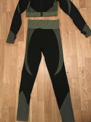 Svart och grön träningsoverall set - Snyggt tvådelat träningsset med croppad zip-jacka och höga tights i svart med gröna detaljer och ränder. Stretchigt material som sitter tight och följer kroppen, perfekt för gymmet eller löpning. Jackan har hög krage och dragkedja framtill.