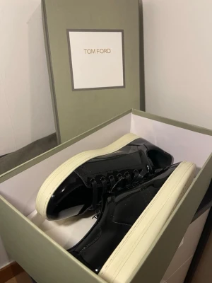  Tom Ford skor - Snygga svarta sneakers från Tom Ford i glansigt lackmaterial med vita, tjocka sulor. Klassisk låg modell med svarta skosnören och diskret perforerat mönster på sidan. Perfekta för dig som vill ha en clean och exklusiv look.