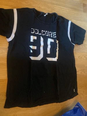 Svart t-shirt med tryck och vita detaljer - Cool svart t-shirt med stort vitt tryck '90' och texten 'SOLOCORE' på bröstet. T-shirten har korta ärmar med vita ränder längs axlarna för en sportig look. Perfekt för dig som gillar streetwear och enkel stil.