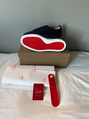 Marinblå Christian Louboutin sneakers - Säljer ett par marinblå sneakers från Christian Louboutin i mocka med ikoniska röda sulor och svarta nitar på tån. Skorna har vita sulor och klassisk låg profil. Grymt snygga detaljer och exklusiv känsla. Perfekta för dig som vill sticka ut med stil.