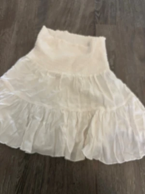 Vit volangkjol med bred midja - Säljer en söt vit kjol med bred smock i midjan och flera lager volanger. Kjolen har en luftig och flowy känsla, perfekt för varma dagar. Passar dig som gillar romantisk och bohemisk stil.