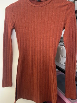 Brun/orange stickad tröja - Rostbrun stickad klänning/tröja med mönster och lång ärm. Modellen är kroppsnära med slits i sidan. Perfekt för dig som gillar en enkel men snygg look. Passar perfekt över kostymbyxor/jeans och ger figurnära passform. Använd ett par gånger men i mycket god skick!