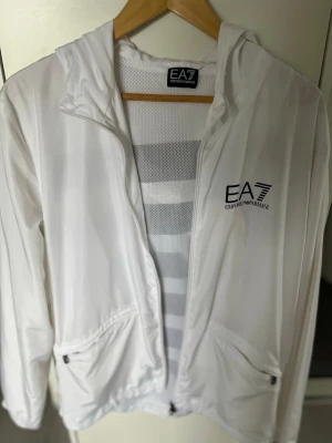 Vit vindjacka från Emporio Armani EA7 - Snygg vit vindjacka från Emporio Armani EA7 med dragkedja framtill, huva och två fickor med dragkedja. Jackan har meshfoder för extra ventilation och en stilren logga på bröstet. Perfekt för en sportig och trendig look.