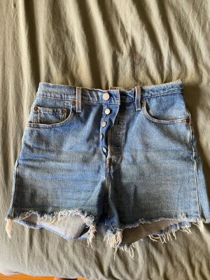 Levi's blå denimshorts hög midja - Säljer ett par klassiska blå denimshorts från Levi's med hög midja och råa, fransiga benslut. Shortsen har fem fickor, knappgylf och den ikoniska Levi's-lappen bak. Perfekta för sommaren och ger en avslappnad vintagekänsla.