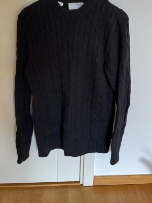 Svart kabelstickad tröja Selected - Kabelstickad svart tröja från Selected med rund halsringning och ribbade muddar vid ärmslut och nederkant. Klassisk modell med lång ärm och snygg struktur i materialet. Perfekt för dig som gillar stilrena och tidlösa plagg.