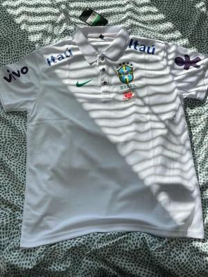 Vit Brasil Nike pikétröja - Säljer en vit pikétröja från Nike med Brasilien fotbollslandslagets emblem på bröstet. Tröjan har tryckta loggor från sponsorer som Itaú och Vivo, samt grönt Nike-märke. Klassisk krage med knappar och korta ärmar. Perfekt för dig som älskar fotboll och vill ha en clean look.