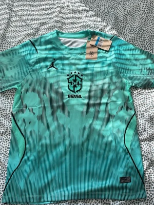 Turkos Brasil tränings t-shirt Nike - Snygg turkos tränings t-shirt från Nike med Brasil-logga och svarta detaljer. Tröjan har korta ärmar, rund hals och ett sportigt mönster i svart. Tillverkad i lätt och ventilerande material, perfekt för fotboll eller träning.