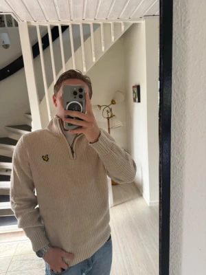 Beige ribbad tröja från Lyle & Scott - Säljer en beige ribbad tröja från Lyle & Scott med halv dragkedja och den klassiska gula loggan på bröstet. Tröjan har hög krage och är tillverkad i ett mjukt stickat material. Perfekt för en avslappnad och stilren look. Origan pris runt 1500kr