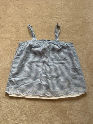Blåvit linnetopp från GAP - Söt linnetopp med smala blå och vita ränder, justerbara axelband och broderade blommor längs fållen. Toppen har en luftig passform och är perfekt för varma dagar. Materialet känns lätt och bekvämt mot huden. Köpte i Los Angeles från GAP ✨🩵