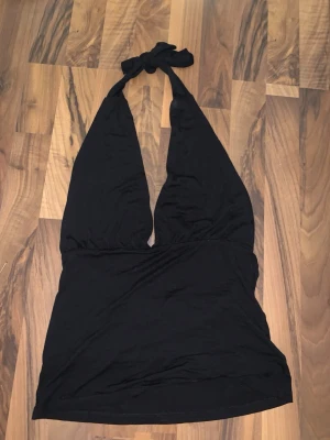 Svart halterneck topp med knyt - Snygg svart halterneck topp med knyt i nacken och djup v-ringning. Toppen är ärmlös och har en figurnära passform som framhäver axlarna. Tillverkad i mjukt bomullsmaterial, perfekt för en trendig och avslappnad stil.