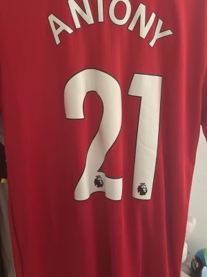 Manchester United tröja Antony 21 - Röd Manchester United matchtröja från Adidas med spelarnamnet Antony och nummer 21 på ryggen. Tröjan har klubbmärke, Adidas-logga och TeamViewer som sponsor framtill. Perfekt för fotbollsfans som vill visa sitt stöd på läktaren eller planen.