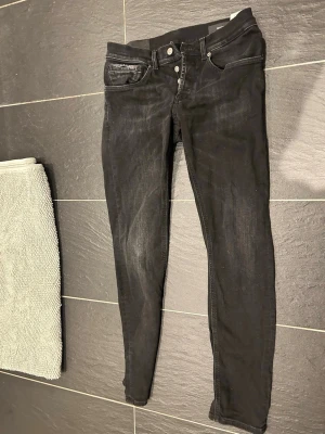 Svarta skinny jeans från dondup  - Säljer ett par svarta skinny jeans från dondup med diskreta slitningar och klassisk femficksmodell. Jeansen har silverfärgade knappar och detaljer samt en liten D-logga på ena bakfickan. Perfekta för en stilren och avslappnad look.