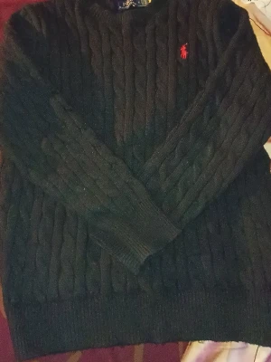 Svart kabelstickad tröja Polo Ralph Lauren - Kabelstickad svart tröja från Polo Ralph Lauren med rund halsringning och den klassiska röda logotypen broderad på bröstet. Tröjan har långa ärmar och ribbade muddar. Perfekt för dig som gillar stilrena och tidlösa plagg.