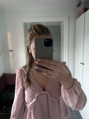 Ljusrosa blus med prickigt mönster - Söt ljusrosa blus med v-ringning och volangdetaljer. Blusen har ett prickigt mönster i tyget och långa ärmar med resår vid ärmslut. Materialet är lätt och transparent, perfekt för att skapa en romantisk look.