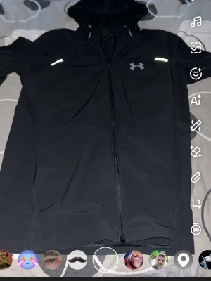Svart vindjacka från Under Armour - Snygg svart vindjacka från Under Armour med huva och hel dragkedja framtill. Jackan har reflekterande detaljer på bröstet och stor logga på ryggen. Tillverkad i lätt och smidigt material som passar perfekt för träning eller när det blåser ute.