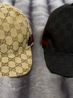 Gucci kepsar med GG-mönster - Två snygga kepsar från Gucci med klassiskt GG-mönster. En i beige med bruna detaljer och en i svart, båda med den ikoniska gröna och röda randen på sidan. Perfekt accessoar för att lyfta din outfit och visa din stil.
