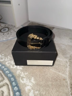 Versace jeans couture svart bälte med guldspänne - Snyggt svart äkta skärp från Versace jeans couture med ett detaljerat guldspänne som har barockinspirerade mönster. Skärpet är gjort i skinn och ger en lyxig känsla till din outfit. Perfekt accessoar för att lyfta vilken look som helst.