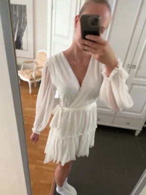 Vit klänning  - Säljer en superfin vit omlottklänning med långa, transparenta ärmar och volangdetaljer. Klänningen har v-ringning, knyt i midjan. Funkar super bra för både student, skolavslutning, midsommar och konfirmation. 