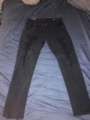 Dondup jeans med slitningar - Säljer ett par svarta jeans med raka ben och slitna detaljer framtill. Jeansen har klassisk femficksmodell och knappgylf. Materialet är denim i bomull och de har en avslappnad passform som passar till många olika stilar.
