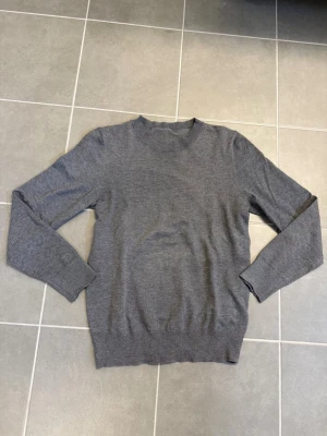 Grå stickad tröja med rund hals - En enkel och stilren grå stickad tröja med rund halsringning och långa ärmar. Tröjan har ribbade muddar vid ärmslut och nederkant. Perfekt att matcha med jeans eller kjol för en clean look.