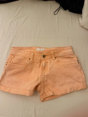 Lågmidjade shorts i aprikosfärg - Snygga aprikosfärgade jeansshorts med klassisk femficksdesign, knapp och dragkedja framtill samt hällor för skärp. Perfekta för varma dagar och har en enkel, stilren look.