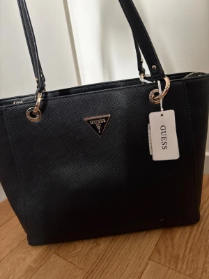 Svart handväska från Guess - Stilren svart handväska från Guess med guldfärgade metalldetaljer och klassisk trekantig logga framtill. Väskan har två långa axelband och är tillverkad i ett strukturerat konstlädermaterial. Perfekt för dig som gillar en elegant och tidlös look.