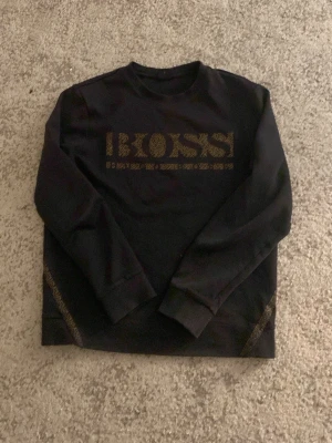 Svart sweatshirt från BOSS - Cool svart sweatshirt från BOSS med guldiga detaljer och stort BOSS-tryck framtill. Tröjan har rund halsringning och långa ärmar. Perfekt för dig som gillar stilrena och trendiga plagg med en exklusiv touch.