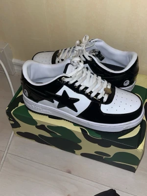 BAPE Sta svartvita sneakers - Säljer ett par BAPE Star sneakers i svart och vitt med glansiga paneler och den ikoniska stjärnan på sidan. Skorna har vita snören, perforerad tåbox och en klassisk låg siluett. Kommer i originalkartong med camomönster. Perfekta för dig som gillar streetwear.