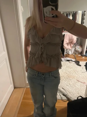 Beige volangtopp med knyt - Supersöt beige topp med volanger och knyt framtill. Toppen är ärmlös och har en croppad passform med v-ringning. Perfekt för dig som gillar romantiska detaljer och en luftig känsla.