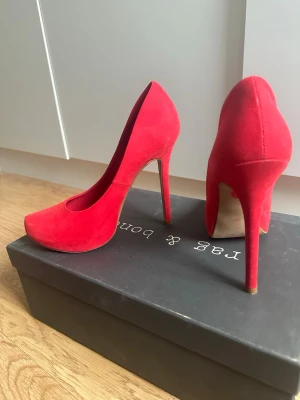 Röda pumps - Snygga röda pumps från Jas & Bond i mjuk mocka med klassisk spetsig tå och hög smal klack. Skorna har en elegant siluett och är perfekta för att lyfta vilken outfit som helst. Materialet ger en lyxig känsla och färgen sticker verkligen ut.
