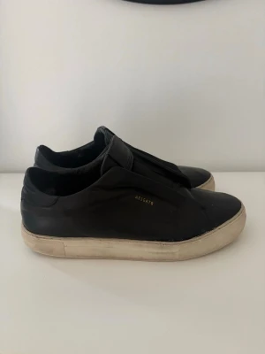 Svarta sneakers från Axel Arigato - Säljer ett par svarta sneakers från Axel Arigato med minimalistisk design och diskret logga i guld på sidan. Skorna är tillverkade i läder och har en vit, robust gummisula. Perfekt för dig som gillar stilrena och trendiga skor.