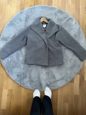 Grå ullkappa med bred krage - Stilren grå kappa i ullmix med bred, markerad krage och tre knappar framtill. Kortare modell som ger en modern siluett. Perfekt för dig som vill ha en enkel men trendig look till hösten eller vintern.