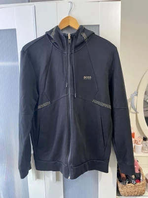 Hugo Boss Hoodie / kofta / luvtröja med zip - En mycket fin Hugo Boss huvtröja med dragkedja/ kofta med zip Svart färg med guldiga detaljer  Storlek M på lappen I nyskick Skicka meddelande vid några funderingar Kolla gärna även in mina andra annonser