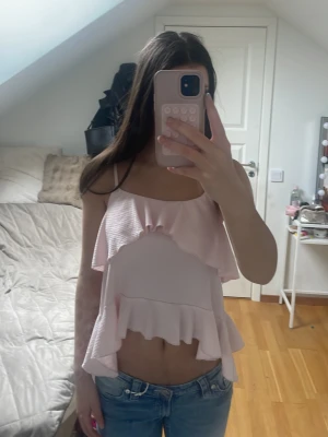 Ljusrosa volang topp med smala band - Supersöt ljusrosa topp med volangdetaljer och smala axelband. Toppen är croppad och har en luftig passform, perfekt för varma dagar. Materialet känns mjukt och lätt mot huden. Passar dig som gillar en feminin och trendig stil.