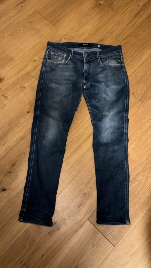 Mörkblå Replay jeans med slitningar - Snygga mörkblå jeans från Replay med klassisk femficksdesign och diskreta slitningar framtill. Jeansen har raka ben och kontrastsömmar, samt Replay-logga på bakfickan. Perfekta för en avslappnad och trendig look.