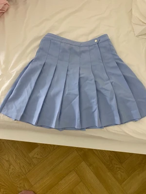 Ljusblå plisserad kortkjol - Superfin ljusblå kortkjol med plisserad design och knappdetalj i midjan. Kjolen har en klassisk tennisstil och är perfekt för dig som gillar en söt och sportig look. Passar till många olika outfits och är lätt att matcha.