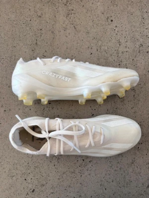 Vita Adidas Crazyfast fotbollsskor - Säljer ett par vita Adidas Crazyfast fotbollsskor med snörning och gummidobbar. Skorna har en modern, strömlinjeformad design och är tillverkade i syntetmaterial för lätt vikt och snabbhet på planen. Perfekta för dig som vill ha stilrena och snabba skor till matchen. Var för små så knappt använda.