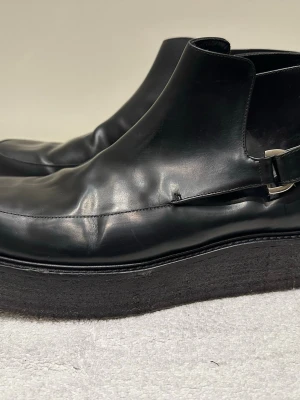 Svarta Whyred läderboots - Fina svarta läderboots från Whyred i bra skick. Stilren design utan snörning med spänne på sidan. Knappt använda. Aningen smal design. Säljer då de tyvärr inte passar mina bredare fötter.