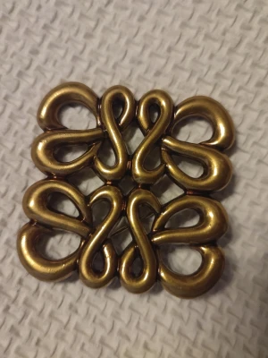 Vintage guldfärgat brosch i metall - Unikt vintage brosch i guldfärgad metall med dubbla, snirkliga former som bildar ett symmetriskt mönster. Broschen har en fyrkantig design och glänsande yta som ger en cool retrovibe. Perfekt för att piffa upp jackan eller väskan.