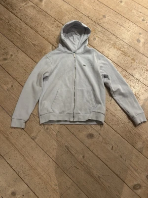 Ljusgrå hoodie med dragkedja Lacoste - Säljer en ljusblå hoodie från Lacoste med dragkedja framtill och klassisk krokodil-logga på bröstet. Hoodien har långa ärmar, fickor fram och är tillverkad i mjuk bomull. Perfekt för en chill och avslappnad stil.