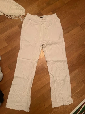 Vita linnebyxor med dragsko - Säljer ett par vita linnebyxor med avslappnad passform och dragsko i midjan. Byxorna har raka ben och sidofickor, perfekta för en chill och stilren look. Materialet är luftigt linne som känns svalt och skönt. 