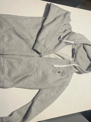 Ralph Lauren hoodie  - Snygg gråmelerad hoodie med dragkedja från Polo Ralph Lauren. Klassisk huva med vita snören, två stora fickor fram och ikoniska mörkblå loggan broderad på bröstet. Mjuk bomullsblandning som är skön att ha på sig och passar perfekt till jeans eller joggers.