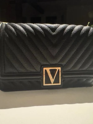Svart Valentino handväska - Snygg svart handväska från Victoria secret med quiltad design och guldigt V-emblem på framsidan. Väskan är tillverkad i läder och har en stilren, rektangulär form. Perfekt för dig som gillar klassiska och trendiga accessoarer. Väskan är köpt i New York men är knappt använd